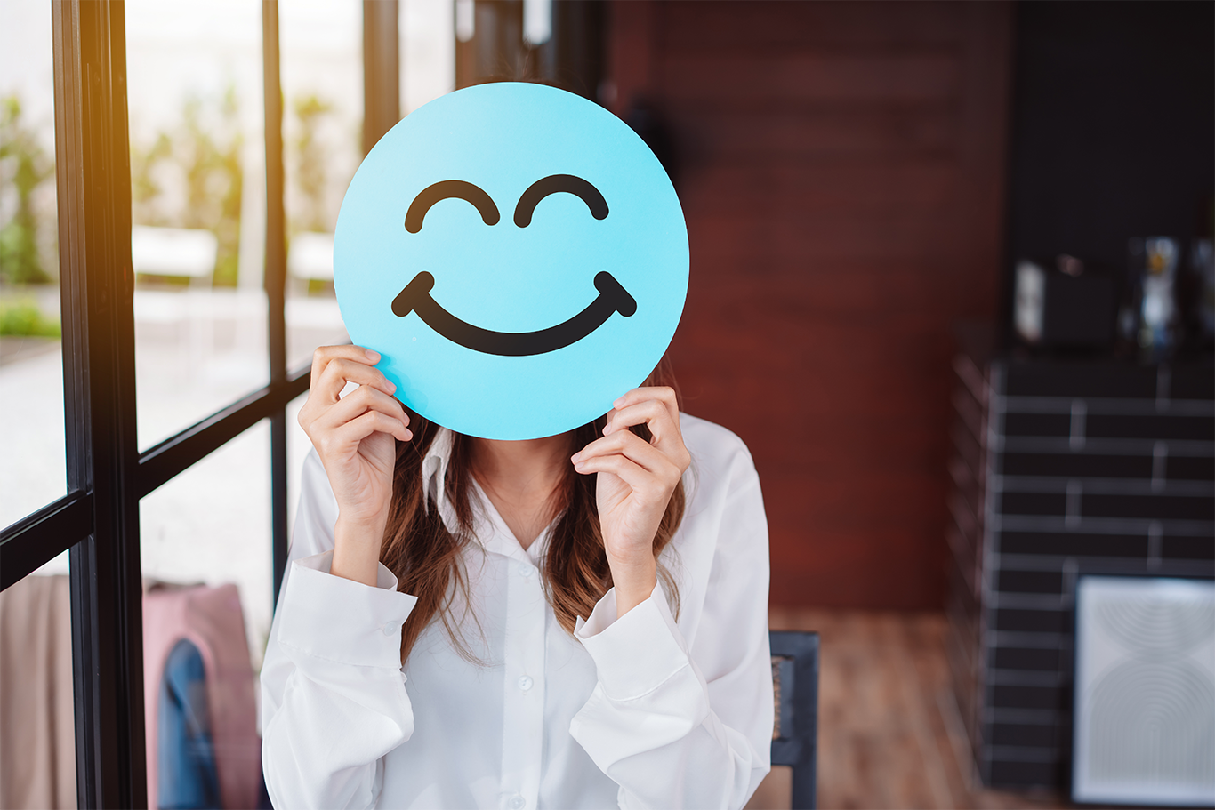 Image of a woman in a professional work setting holding a smiley face icon in front of her face to support good employee article