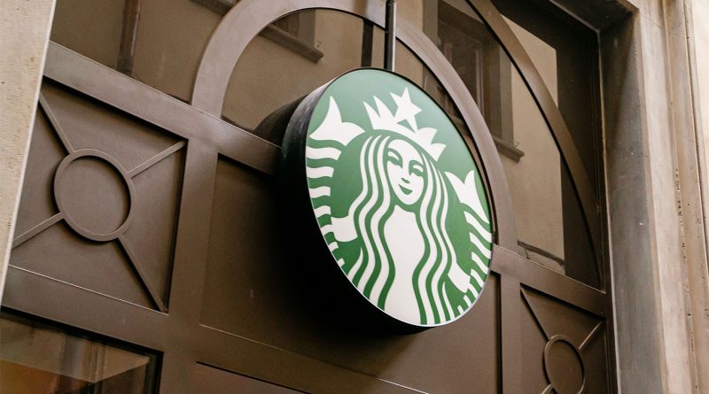 A Starbucks barista prepares a protein latte beside a digital order screen showing voice command features.