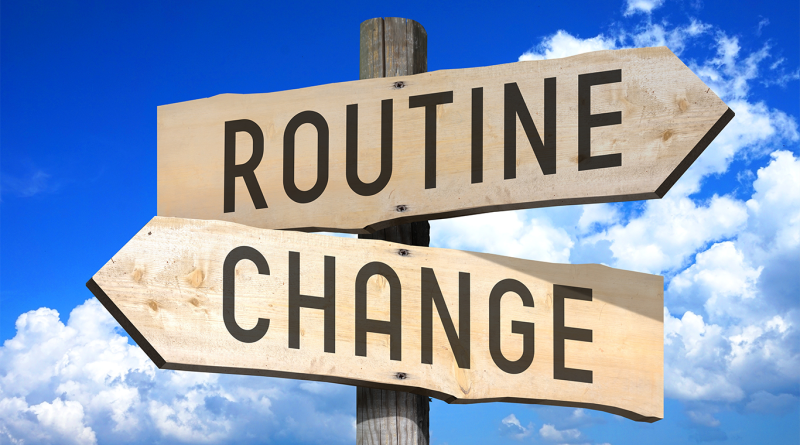 Wooden signpost with two arrow signs reading “ROUTINE” (pointing right) and “CHANGE” (pointing left) against a bright blue sky with clouds.