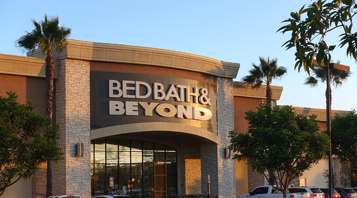 Exterior of a Bed Bath & Beyond store with large signage above the entrance, framed by palm trees and a parking lot at sunset.