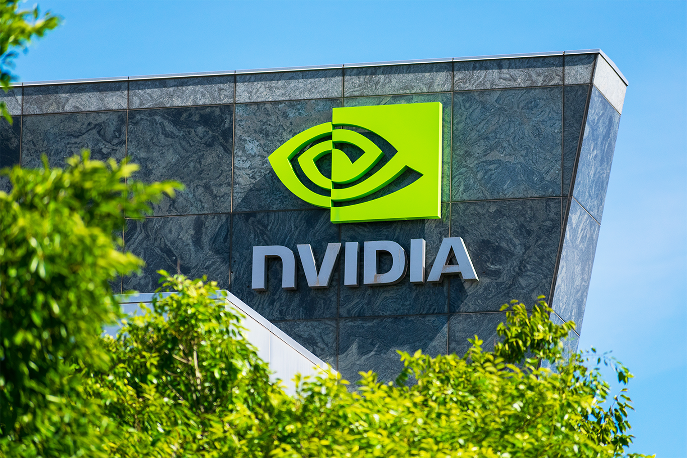 NVIDIA logo and signage on the exterior of a modern building, partially framed by green trees under a clear blue sky.