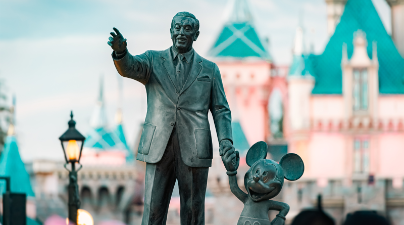 Statue of Walt Disney holding Mickey Mouse’s hand in front of a Disney theme park castle, with teal rooftops and soft evening light in the background.