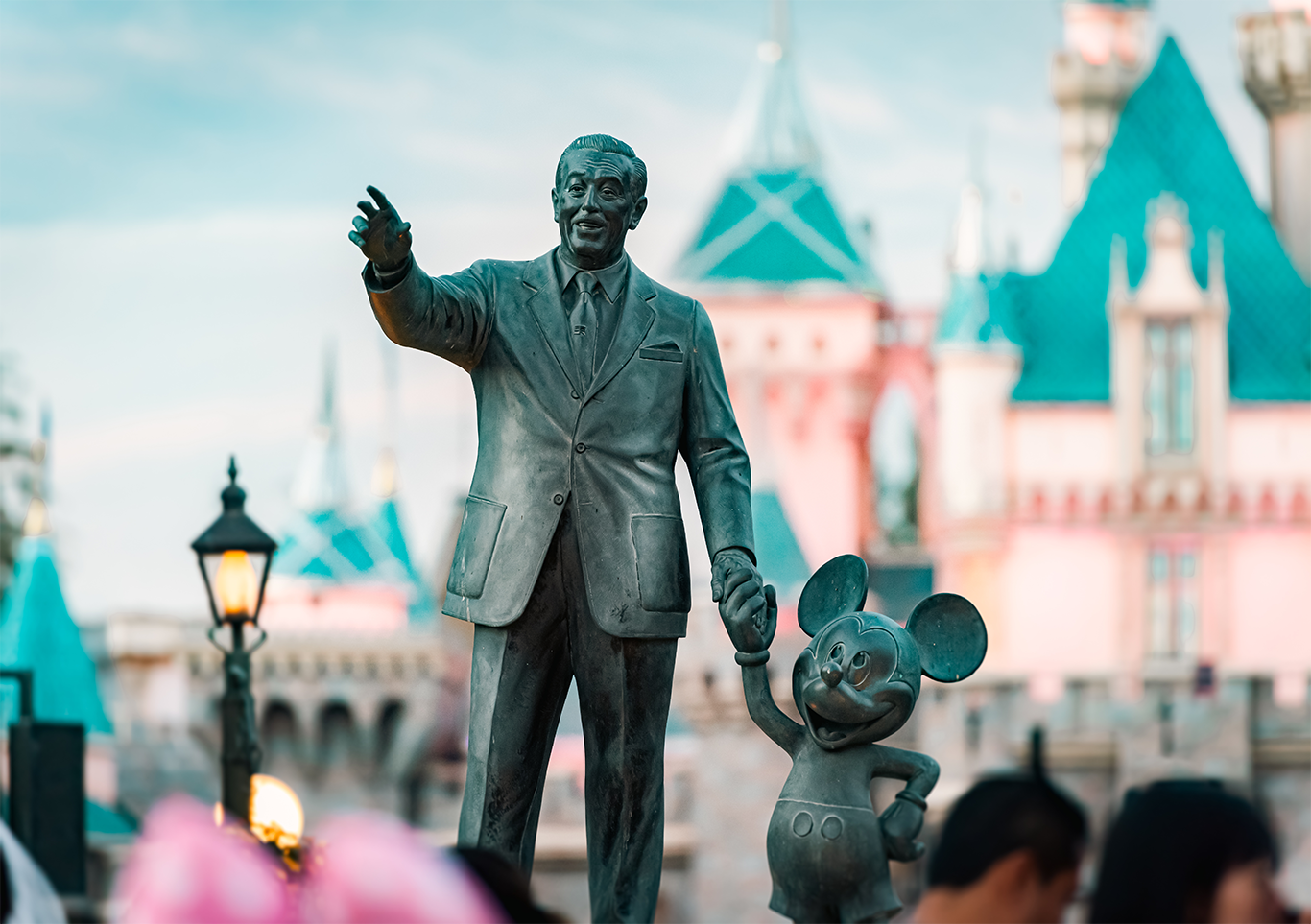 Statue of Walt Disney holding Mickey Mouse’s hand in front of a Disney theme park castle, with teal rooftops and soft evening light in the background.