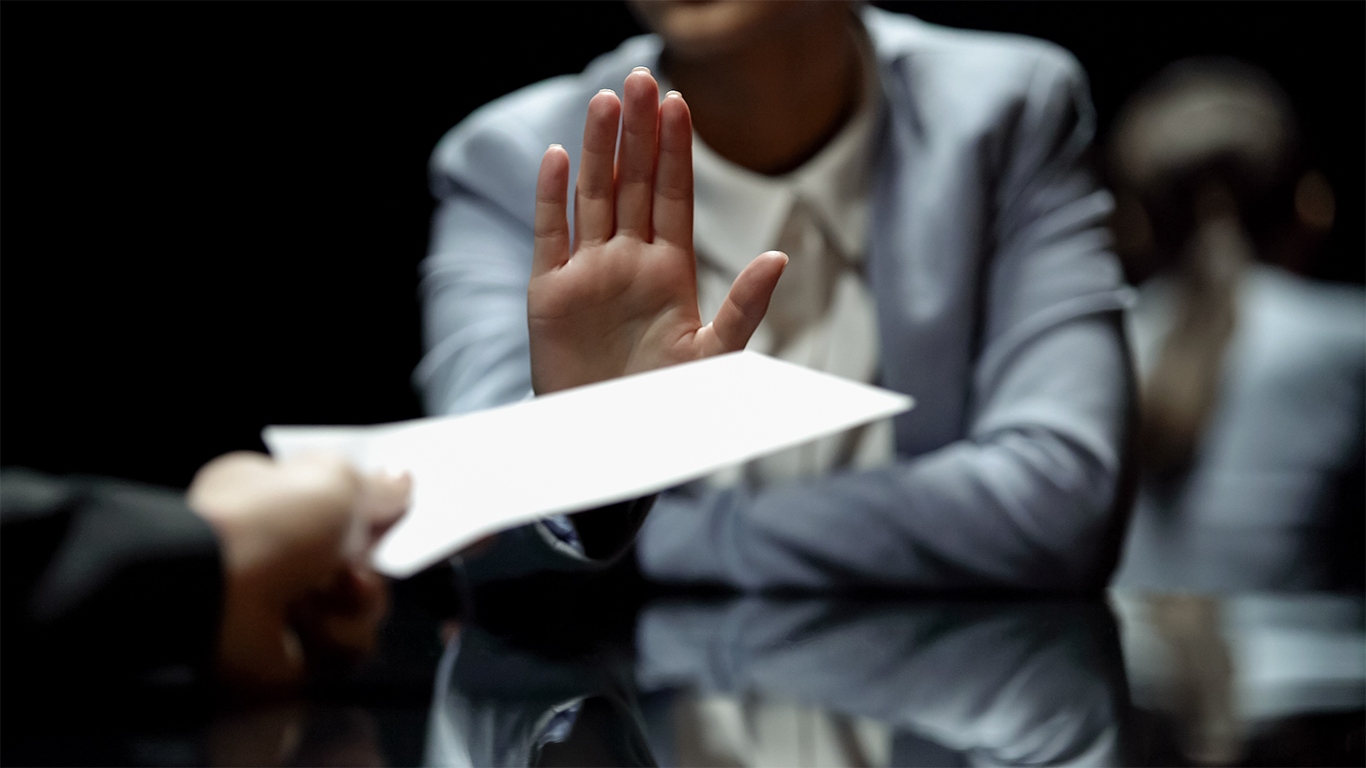 Professional woman in a business suit raising her hand to refuse or reject a document being offered across a desk during a meeting, symbolizing turning down a job offer or salary proposal.